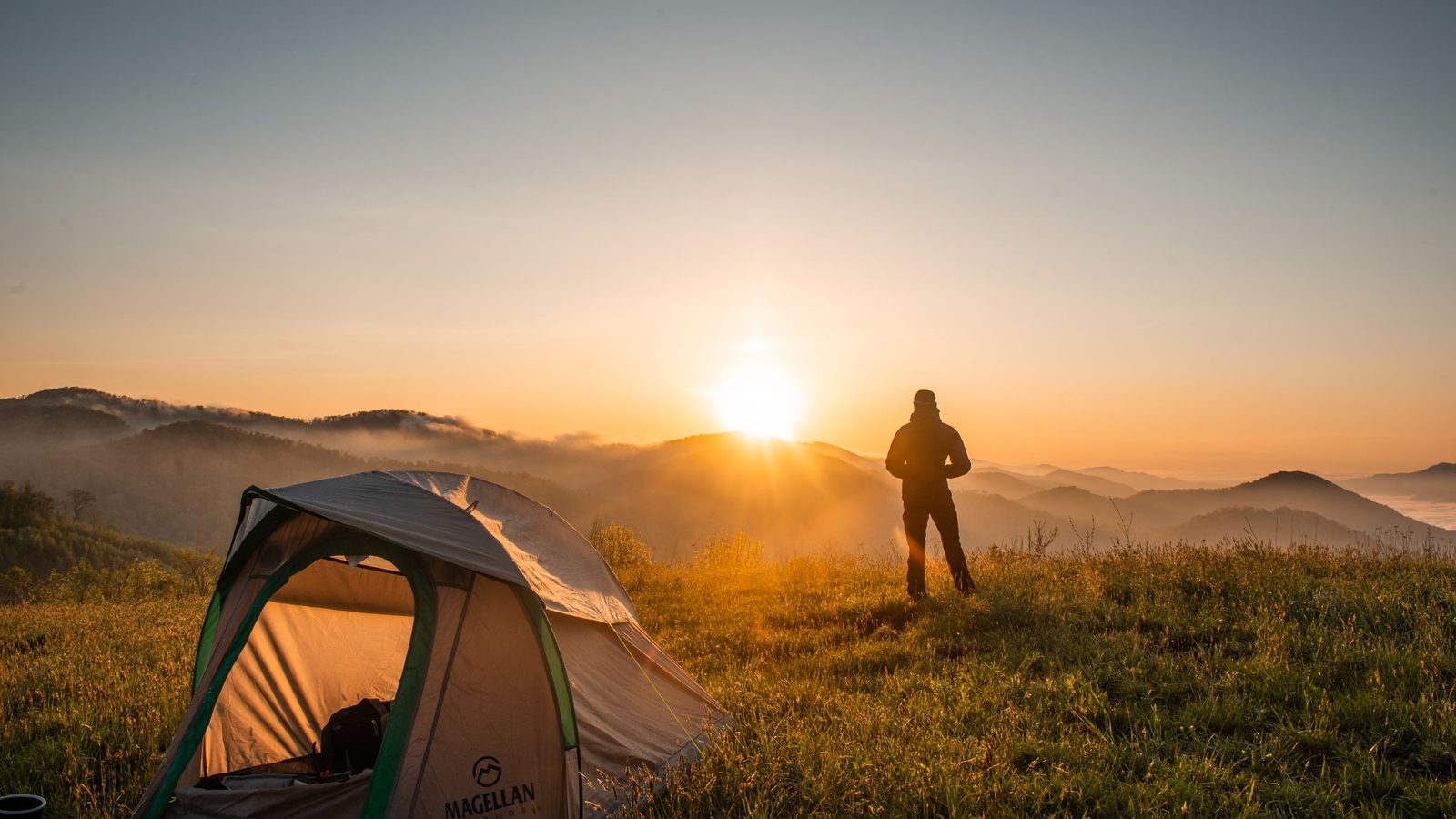A person standing in a field at dawn, watching the sunrise after camping overnight in a tent on a hillside