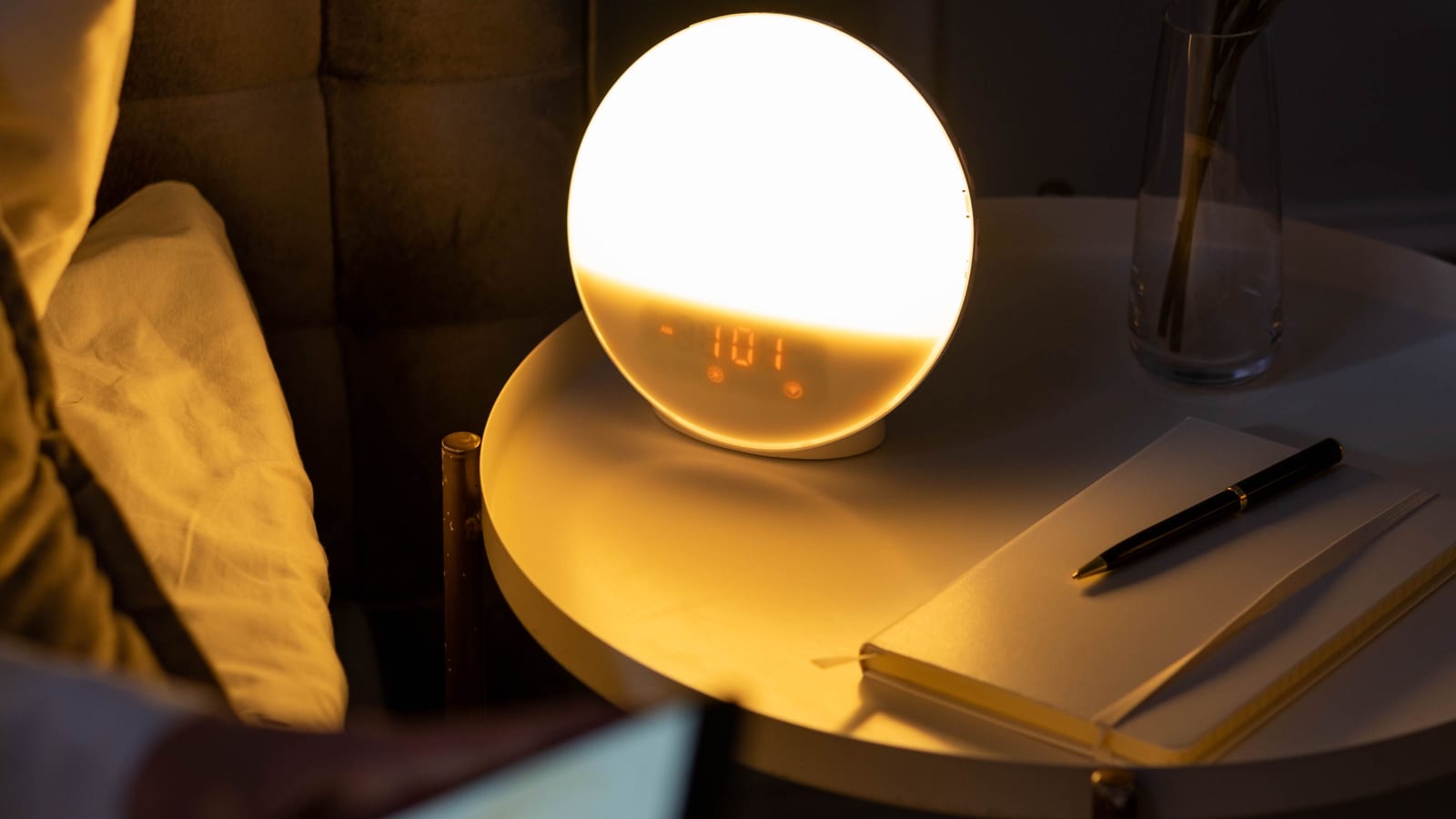 A bedside clock showing 11 PM, with warm lamp light casting long shadows across a pillow and an open alarm app on a phone