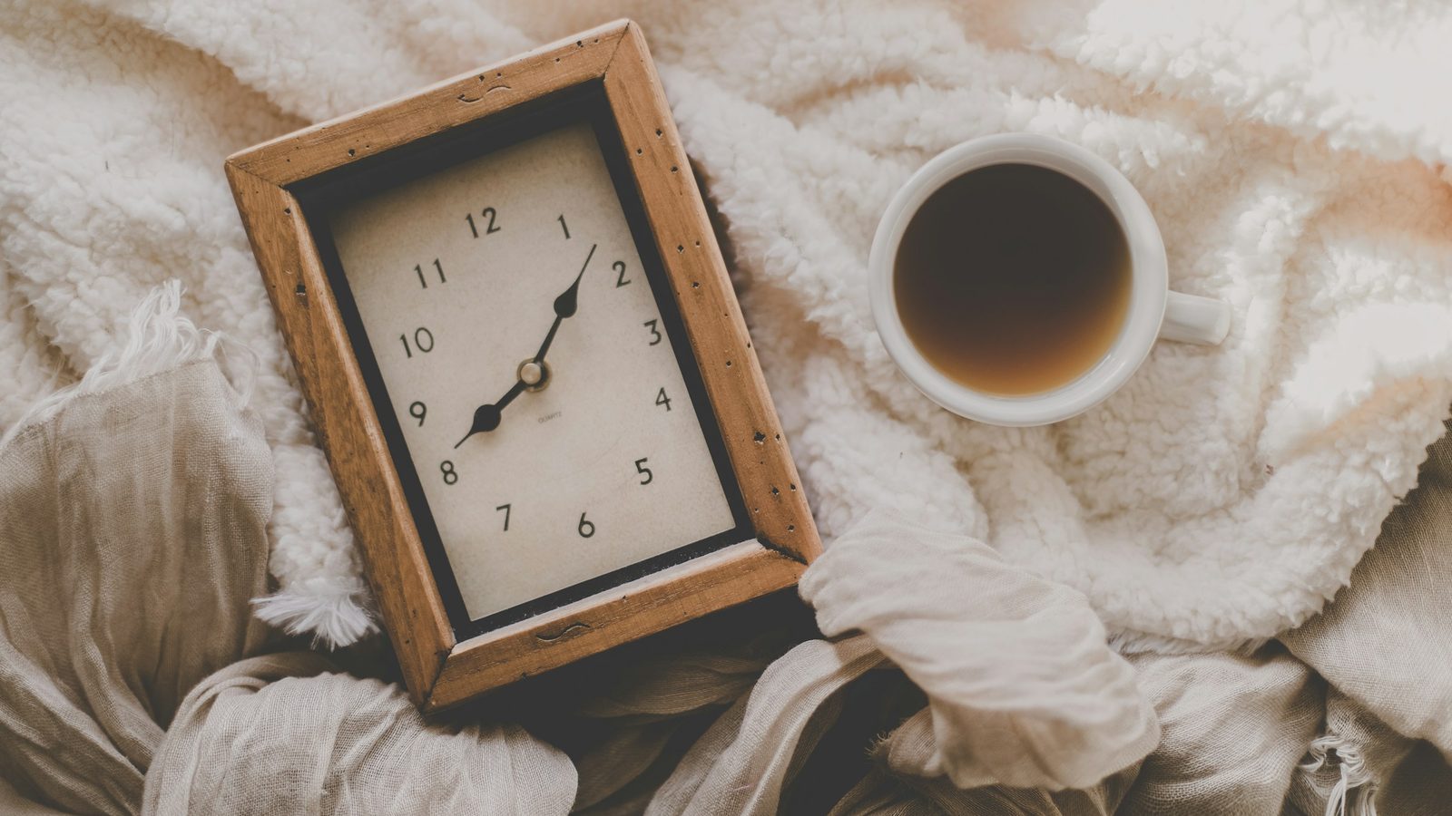 A wooden desk clock reading a little after eight next to a full cup of black coffee on a cream blanket