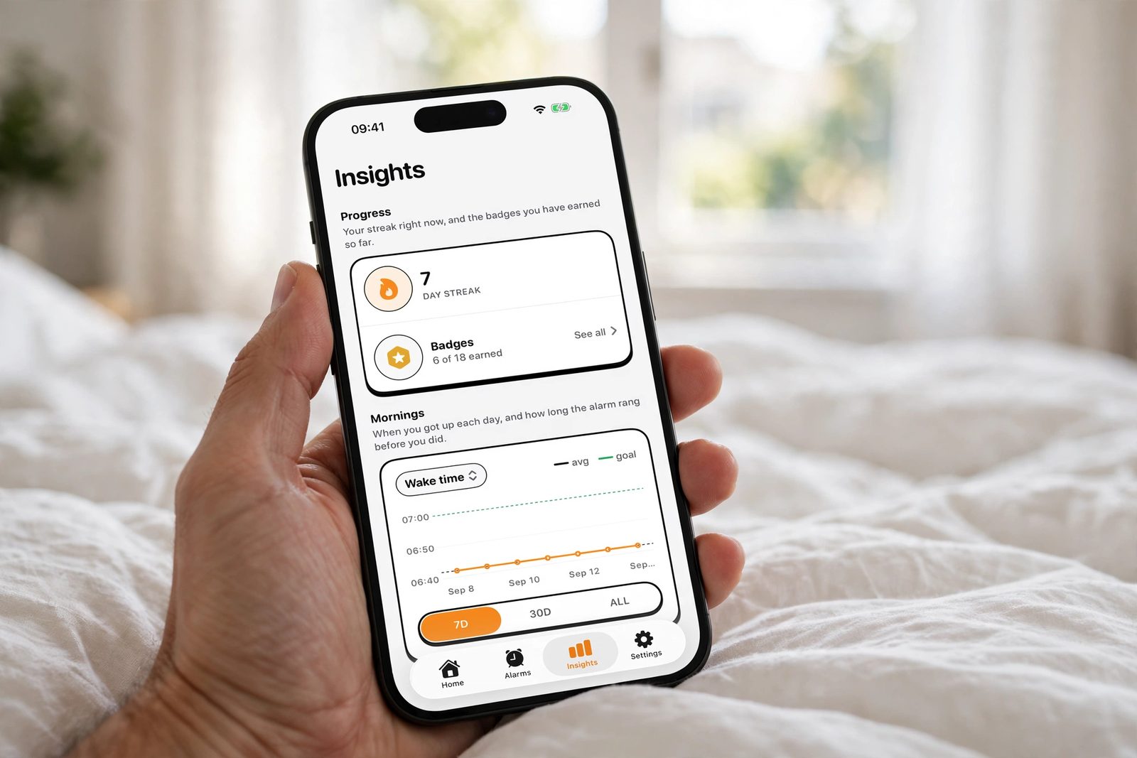A hand holding an iPhone over a bed in morning light, showing NoNap's insights dashboard with wake-up time trends and streak consistency over recent days