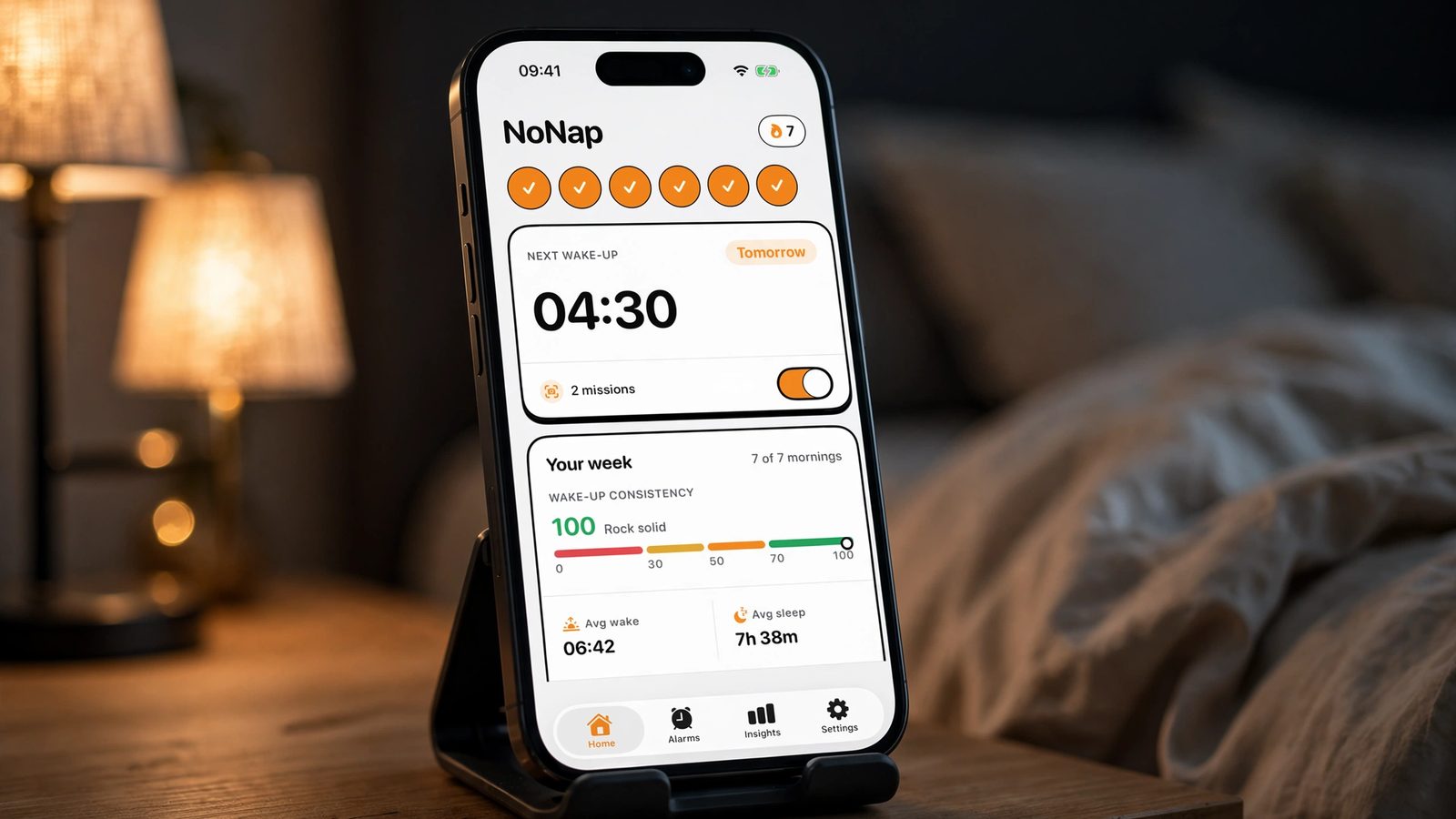 An iPhone on a stand on a bedside table before dawn, showing the NoNap home screen with a 04:30 next wake-up and a seven-day streak