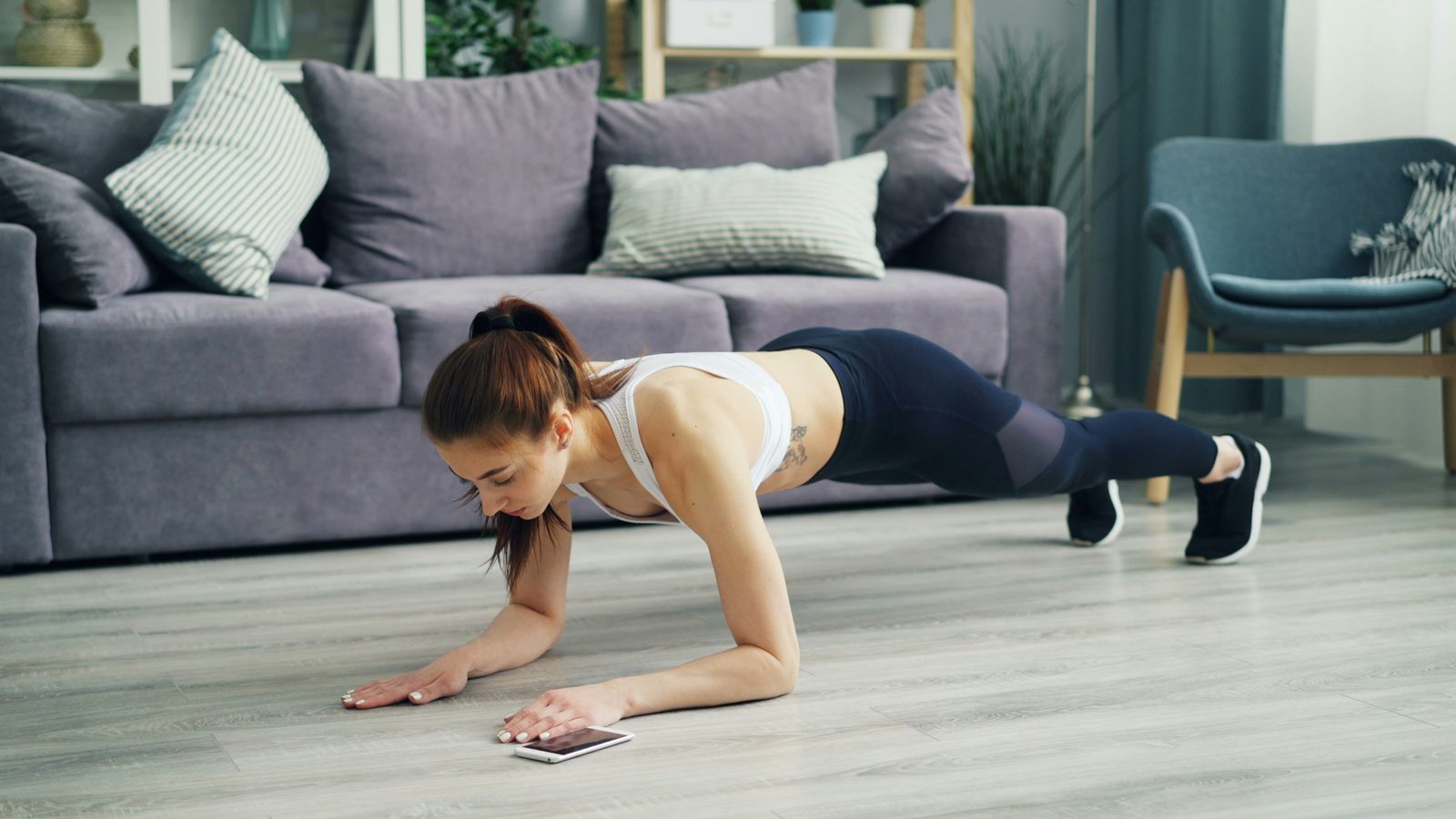 A woman doing a push-up on a wooden floor in a bright living room with plants and a sofa in the background