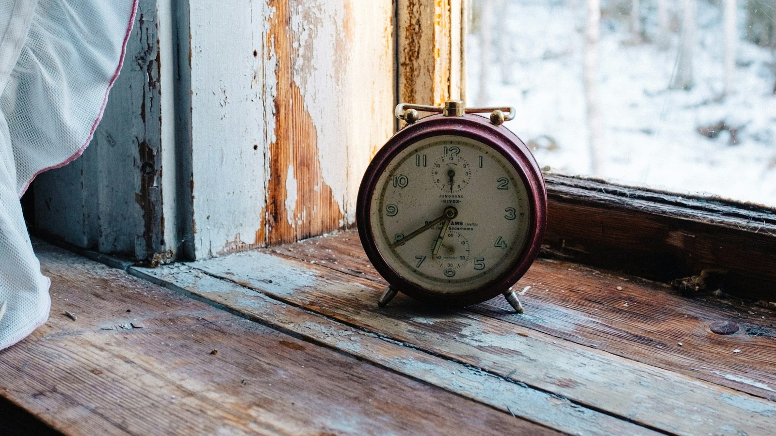 A worn red alarm clock sitting on a windowsill, with bright snowy daylight visible through the window behind it