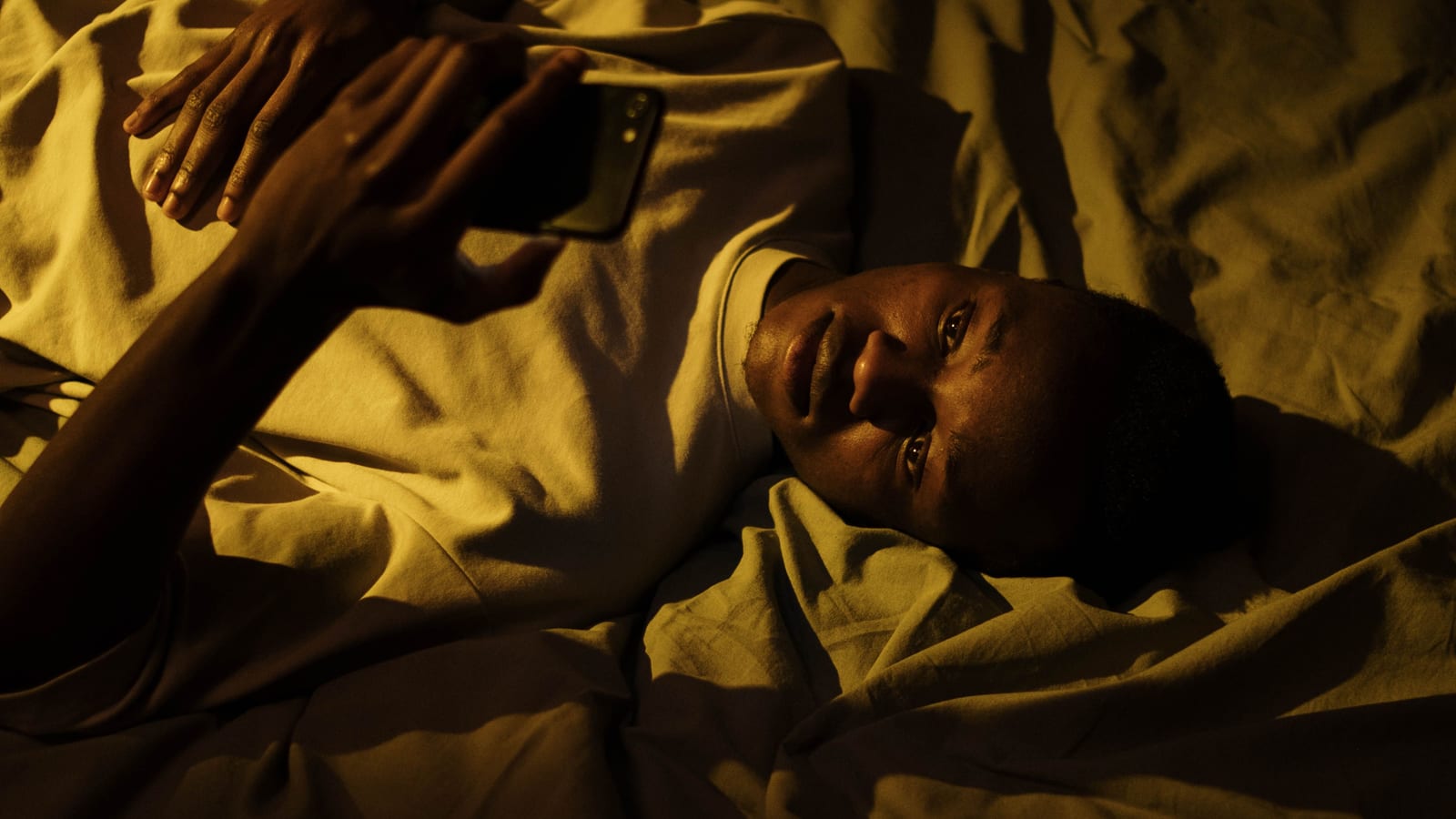 An iPhone on a nightstand glowing in a dark bedroom at night, with blue light illuminating the face of a person in bed