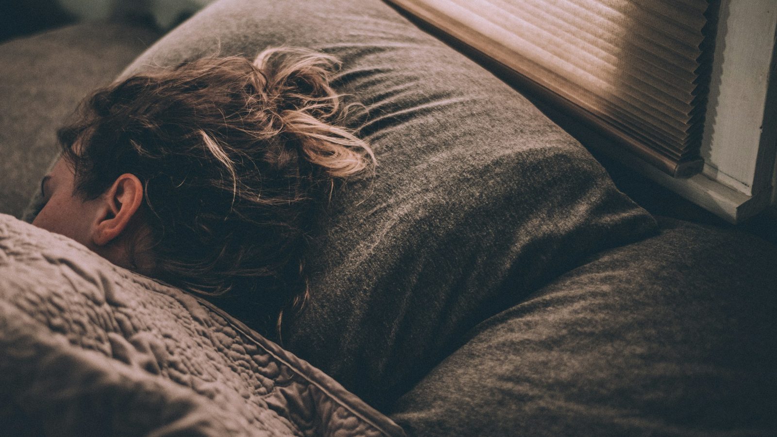 A person sleeping in late under warm blankets as morning light comes through window blinds